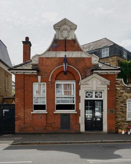 Charmant petit bâtiment bas en brique rouge et pierre blanche présentant un pignon central en haut duquel on peut lire l’année de construction, 1906. Une grande fenêtre centrale surmontée de briques en arc plein cintre est flanquée à gauche d’une plus petite fenêtre rectangulaire plus modeste et à droite d’une porte au tympan blanc triangulaire.