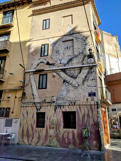 A four-story building in Valencia, Spain, features a large-scale mural of a whimsical cat-like creature on its side. The mural is rendered in shades of gray and white, creating a striking contrast against the building's beige facade. The creature has large eyes, a long snout, and holds its tail that has caught fire. The cat looks desperate or resigned. Below the mural, painted flames rise from the base of the building, adding a dramatic touch. The building has several windows and balconies, and the street level features a doorway and a small table and chairs.