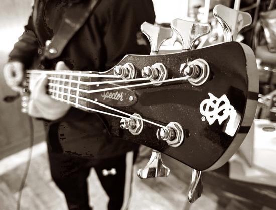 A close-up in black-and-white view of the headstock of a bass guitar. The brand name "Spector" is visible in a cursive font. A distinctive logo is also visible on the headstock. The tuning pegs are clearly shown, with the strings tightly wound around them. The background is blurred, but a person is holding the bass guitar, emphasizing the headstock and strings.