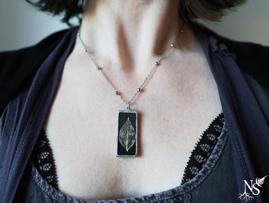 English A woman's neckline with black and dark grey lace top and a simple silver necklace with a stained glass pendant containing real a pressed skeleton leaf on a black background.
❉
Français Un décolleté de femme avec un haut en dentelle noire et gris foncé et un collier simple en argent avec un pendentif en vitrail contenant une véritable feuille pressée dont il ne reste que les nervures sur fond noir.