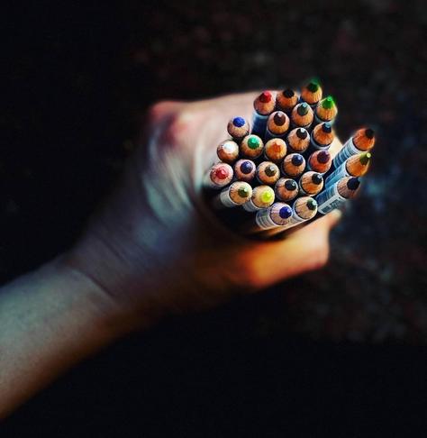 A hand holding a bundle of colored pencils, showcasing a variety of colors at their tips against a dark background.