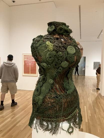 A corset/fitted torso garment on a dress form made with various fibre techniques including weaving and circular formed patterns. The back ground shows the hardwood floored room with a man in shorts and two other patrons in the distance