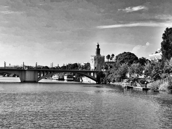 Una vista en blanco y negro del Puente de San Telmo, puente arqueado que cruza el Río Guadalquivir, con la Torre del Oro de Sevilla visible al fondo. El río es tranquilo y refleja el cielo nublado.  Detrás del puente, se ve una línea de árboles y edificios. La Torre del Oro se destaca como un elemento arquitectónico principal en el paisaje.  Una pequeña estructura se encuentra en la orilla del río cerca del margen derecho de la imagen.