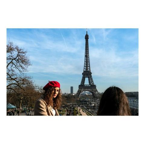 Une femme portant un béret rouge et un manteau beige pose devant la tour Eiffel sous un ciel bleu. Son regard est tourné vers l’objectif, tandis qu’une autre personne, de dos, encadre la scène. L’arrière-plan animé par des promeneurs et l’architecture iconique de Paris évoque une atmosphère typiquement parisienne, entre élégance et effervescence touristique.