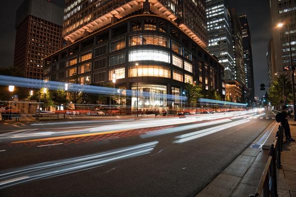 Long-exposure nighttime photograph of Beams Bar in Marunouchi, Tokyo. Bright white, red, and blue light trails from passing traffic streak across the foreground, while the illuminated glass facade of the modern building stands out against the dark urban skyline.