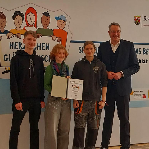 Foto mit Urkunde von drei jungen Menschen der EJN zusammen mit Ministerpräsident Alexander Schweitzer und der Wettbewerbs-Urkunde. Im Hintergrund ein Werbebanner.