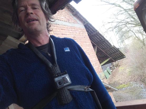 Mann mit blauem Pullover vor Ziegelsteinwand. Auf der Brust sitzt eine Actionkamera, die an einem offensichtlich selbst gebastelten Brustgurtbefestigt ist. Die Grundplatte des Gurts ist ein Stück Fahrradreifen, etwa 15 cm hoch und 6 cm breit. Das Ensemble ist mit Fahrradschauch um den Nacken und die Brust am Körper befestigt. Die Schnalle des Brustgurtes ist das Ventil des Schlauchs.