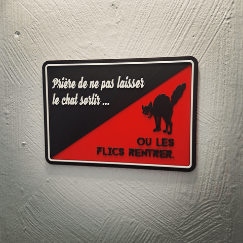 Pancarte imprimé en 3D rouge et noire (reprenant l'esthétique du drapeau de la CNT) avec écrit en haut à gauche "Prière de ne pas laisser le chat sortir..." et en bas à droite "ou les flics rentrer." Avec le chat noir de la CNT.