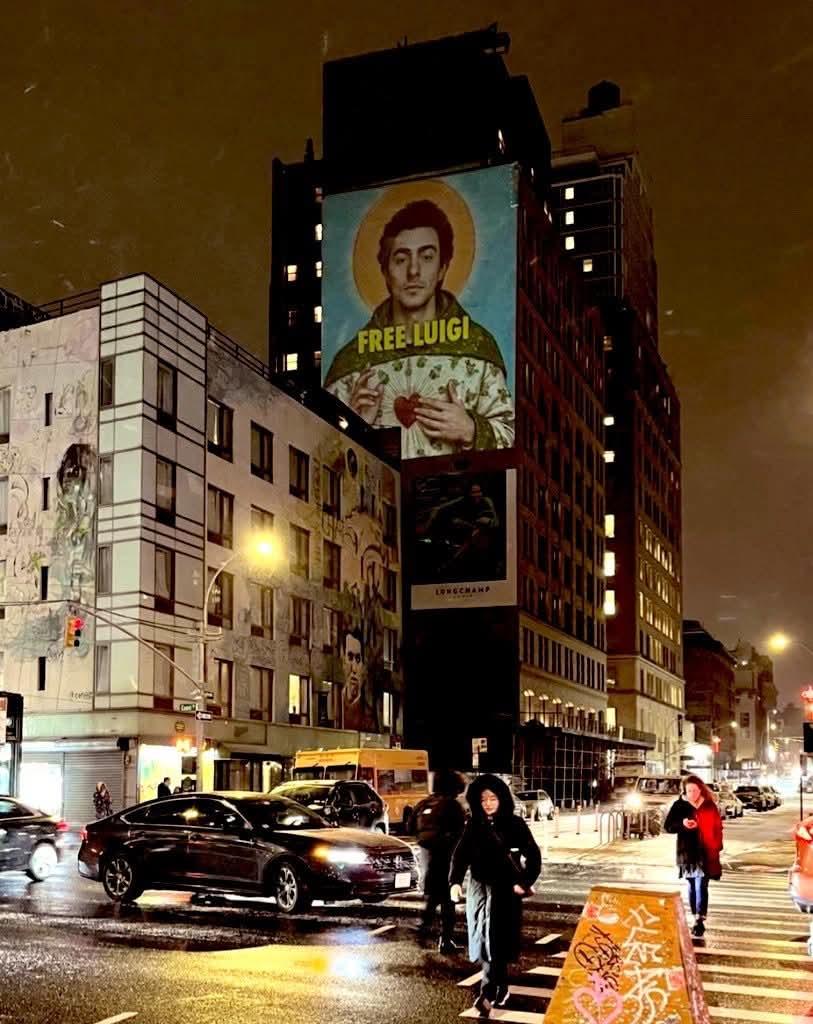 A nighttime cityscape at the intersection of Canal and Lafayette Streets in Lower Manhattan, with wet streets reflecting the glow of streetlights and vehicle headlights. A massive projection on the side of a high-rise building features an image of Luigi Mangione depicted as a saint, holding a flaming heart, with the bold yellow text "FREE LUIGI!" above him. This temporary projection was part of an activist campaign ahead of his court appearance on February 21, 2025. Below, pedestrians in hooded jackets cross the street, while cars and a yellow bus navigate the busy intersection. Another mural decorates the side of a lower building, adding to the urban art scene. A graffiti-covered traffic barrier stands in the foreground, reinforcing the gritty, artistic energy of the area.