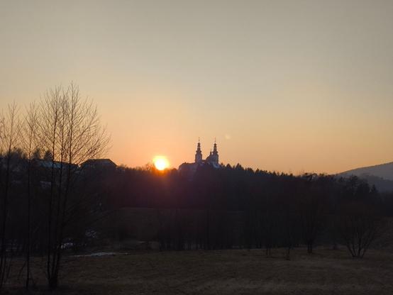 Die Sonne geht direkt neben zwei Kirchturmspitzen unter. Im Vordergrund ein paar kahle Bäume, viel Wiese dazwischen mit grün-braunem Gras.