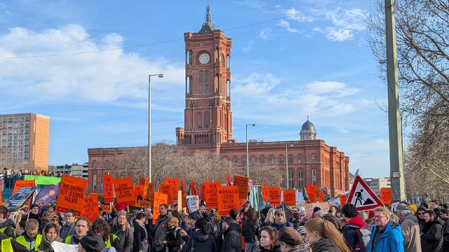 Viele Menschen mit orangenen Schildern stehen auf der Straße. Im Hintergrund ist das rote Rathaus zu sehen.