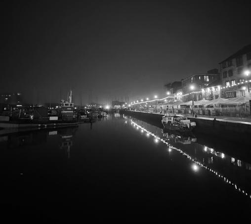 A black and white night time scene of an old waterfront. On the right is a number of bars, with pointed awnings. There's a boat moored along side. A row of lights runs alongside the sea wall. 
These lights are reflected on the water. 
On the left of the scene, in the dark, are an assortment of boats. 
There's a slight mist to the air.