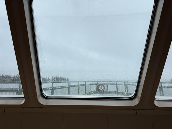 A view through a rain-specked window, showing a misty and overcast landscape outside. The foreground features a portion of a boat's interior, while there are hints of a railing and distant trees visible through the glass.