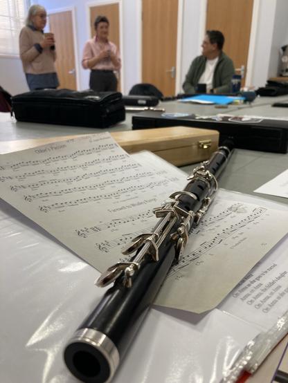A wooden flute lies on a table covered in sheet music. People drink tea and chat in the background.