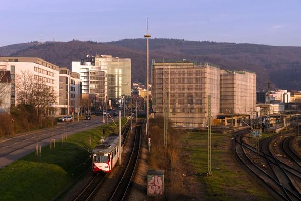 A tram line next to railway tracks passing through office buildings in the evening sun. A red and white 8-axle high-floor tram car, in the usual Düwag design.