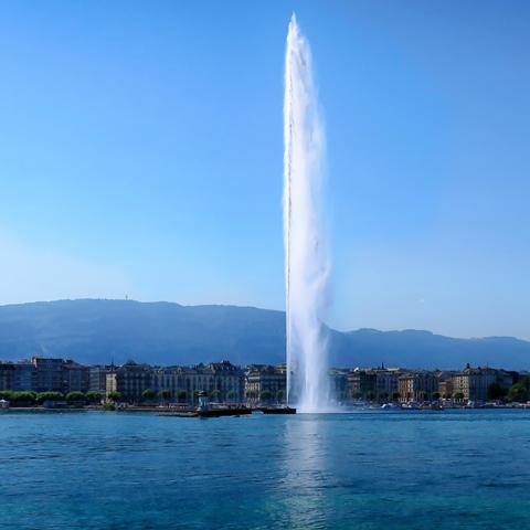 Lake Geneva with the water jet in the center, the lake-side buildings, and the mountain range behind them under a slightly hazy blue sky.