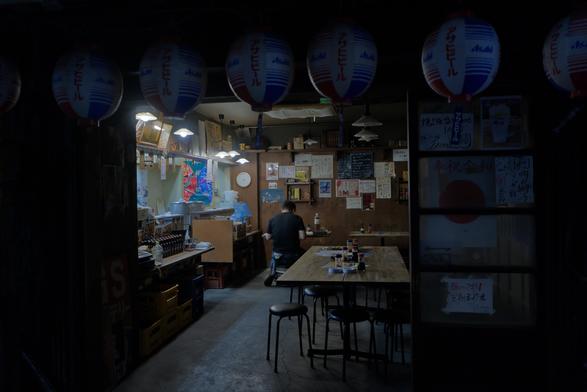 A dimly lit Japanese izakaya with paper lanterns hanging from the ceiling. The space is empty except for the proprietor, who sits at a table with his back to the camera, preparing for the evening. The wooden interior is adorned with handwritten menus, bottles, and small details that create a nostalgic, intimate atmosphere.