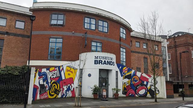 A 1980s building with curved facade and entrance. Painted on the exterior walls are examples of iconic packaging with the trademarks spelt wrong, for example Coolman's Mustard and Waklers crisps. Over the entrance door it says Museum of Brands.