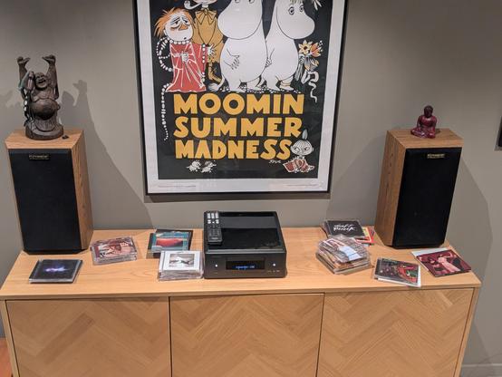 The image presents a scene centered around an audio setup in an apartment.

The focal point is a wooden cabinet adorned with stacks of CDs and a Denon audio player.

Two speakers flank the cabinet, each topped with a statue—one a Laughing Buddha, the other a serene Buddha figure.

Above, a framed poster announces “Moomin Summer Madness”, lending a whimsical touch to the arrangement.