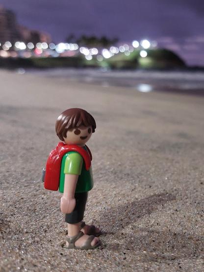 Criança playmobil com mochila vermelha. Na areia da praia, com o mar de frente