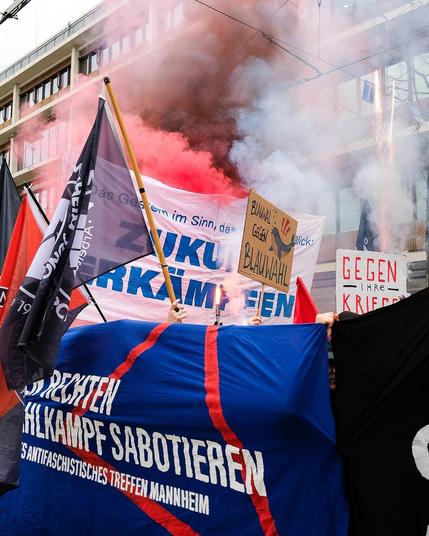Bild mitten in einer dynamischen Demonstration mit vielen Transparenten und Fahnen. Es ist ein Bengalo, Schwarzer und roter Rauch zu sehen.