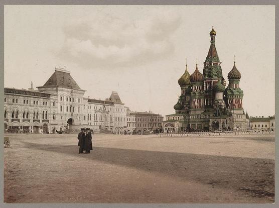The image depicts a historic view of Moscow's Red Square. At the center, we see Saint Basil Cathedral with its iconic onion-shaped domes in vibrant colors such as green, red, and gold against an overcast sky. The architecture surrounding it reflects classical Russian styles.

In the foreground, there are two people walking towards each other on what appears to be a dusty road or square; they're dressed in early 20th-century attire suggesting this photo is from around that time period. Several horse-drawn carriages and horses can also be seen scattered across the scene, which implies it's not just pedestrians but vehicles moving about as well.

The buildings on either side have multiple stories with detailed facades typical of late imperial Russian design. In terms of coloration, this photograph is monochromatic, possibly indicating that colored photography had yet to become widespread or was still in its nascent stages during the era depicted here.