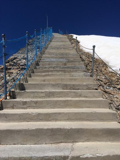 A outside stairway on a mountain, bringing to its summit. We can see about 50 steps until the top. On the right side there is snow and on the background, the sky is clear from any cloud.