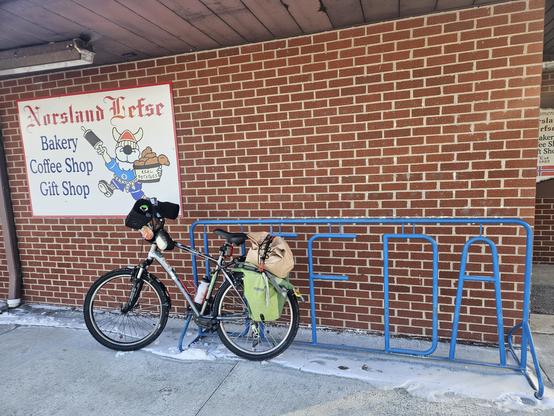 Bicycle with a green bags, a brown bag on top of it's rack is leaning against a bicycle parking rack that says "UFFDA" but the "FDA" are the only visible letters. Behind these us a brick wall. On it is a white sign that says "Norsland Lefse Bakery Coffee Gift Shop" and an illustration of a bearded man in a blue outfit holding a rolling pin raised in the right hand and holding baked goods in the left hand.