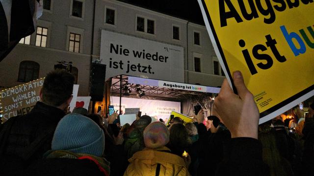Der Himmel ist schwarz, man sieht das Verwaltungsgebäude der Stadt Augsburg. darauf hängt ein großes Schild mit der Aufschrift "nie wieder ist jetzt". Im Vordergrund sieht man eine Hand mit einem Schild "Augsburg ist bunt". etwas weiter hinten ein Schild "demokratisch wählen sonst wird es schmerzhaft" ( die darin enthaltenen Wörter Merz und AfD sind hervorgehoben). viele Menschen mit Schildern stehen mit dem Rücken zur Fotografin vor einer Bühne, auf der gesprochen wird. Auf der Bühne steht "Augsburg ist bunt", "für Vielfalt und Demokratie".