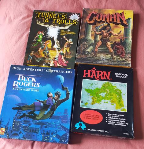 TnT, 5 heroes face off against a huge Troll. One casts take that you fiend, striking the beast which has its arms outstretched. Strong yellows, especially the logo

Conan, he roars as he clenches his fist and holds up his sword. Behind prowls a draconic cat like being. Strong yellows and red dominate the cover.

Buck flies through the air, holding Wilma to him. He wears a leather cap and jacket, she a purple top. Both have jodhpur like trousers. Buck holds a pistol aloft as he looks back and down past his passenger. She almost swoons in his arms. They pass by illuminated buildings in the blue ish cloak of night-time.

Harn, a plain black box with an insert of a map of Harn. An island similar to an enlarged version of the British Isles, smashed together and rotated. It is strongly green isle, with brown marking mountains. Harn is emblazoned in large red letters in the top left.