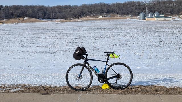Blue and black Roubaix bicycle with Bar Mitts parked on the side of Olmsted County 25 looking North with a mostly snow covered corn field with grain bins in the distance on the right and the bare tree covered Weatherhill behind.