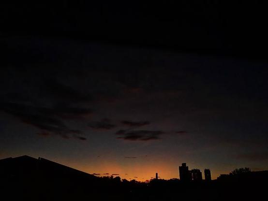 The orange glow of the sun barely over the horizon of rooftops and faraway buildings beginning to illuminate the still dark morning sky. very few clouds in the sky.