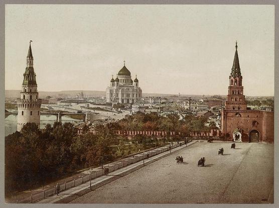 The image depicts a panoramic view of the Moscow Kremlin, with its iconic St. Basil's Cathedral prominently featured in the center background. To the right stands the Spasskaya Tower of the Kremlin Wall, and to the left is the Nikolskaya Tower. The foreground shows Red Square, bustling with activity including horse-drawn carriages moving along a wide street lined by trees. In the distance beyond the Kremlin walls lies the River Moskva winding through Moscow's urban landscape.