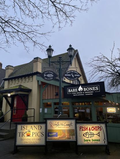A daytime exterior shot of the "Bare Bones Fish House & Smokery" restaurant. The building is a two-story structure with a green-tiled roof, a tan-colored facade, and a red door on the left side. Above the door is the number 4324. A black light post stands in front of the building, with signs hanging from it. The signs read "Bare Bones Fish & Chips" with a fish skeleton logo and "Walk The Coast Footwear Apparel." Above the main entrance of the restaurant is a large black sign that reads "Bare Bones Fish House & Smokery." An "Open" sign hangs in the window.

In the foreground, there are three illuminated signs. The leftmost sign advertises "Island Tropics Tanning Studio," featuring a palm tree and a sunset. The middle sign advertises "Walk the Coast Hikers to High Heels" with a picture of hiking boots and the Blundstone brand. The rightmost sign advertises "Fish & Chips Bare Bones" with a similar fish skeleton logo as the one above. The sky is overcast, and bare tree branches are visible above the building.