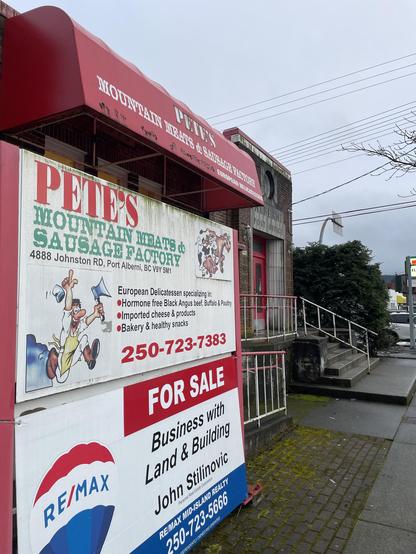 ―
Here is the alt-text description for the image: The image shows the outside of "Pete's Mountain Meats & Sausage Factory". Above the entrance is a red awning with the store's name in white letters. Attached to the building is a sign with a cartoon butcher holding an ax and a cleaver. The sign also lists the store's address, phone number, and products, including hormone-free black Angus beef, imported cheese, and bakery items. Below the store sign is a "For Sale" sign for the business, land, and building, with the RE/MAX logo and the realtor's name and contact information. The building is made of brick and has a small set of stairs leading up to the entrance. A sign above the door says "Alberni Post Office". A small tree is to the right. The sky is overcast.
―