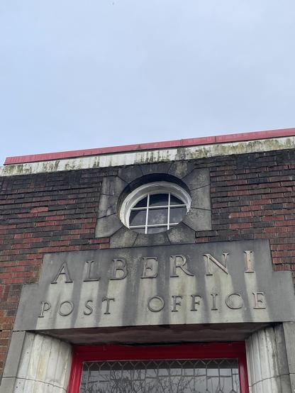 The Alberni Post Office building is shown from below. The building is made of red bricks and is topped with a red roof. The name of the building is in raised lettering. The top line says "ALBERNI" and the second line says "POST OFFICE." Above the lettering is a round window with white bars. The sky is overcast.
―
