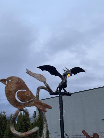 A medium shot shows a rusty metal bird sculpture in the foreground, slightly blurred, and a metal dragon sculpture breathing "fire" atop a pole with lights in the background.

The bird is to the left and is made of brown metal with a large white "C" shape cut out of its side. It is perched on a metal branch-like structure. Behind it are green trees.

In the background, a black metal dragon with wings spread stands on a pole, breathing orange "fire." The pole is decorated with small white lights. Behind the dragon is a plain white building. The sky above is cloudy and gray.
―