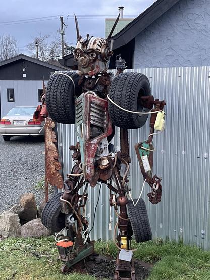 A full shot of a robot sculpture made of scrap metal, car parts, and tires standing on a patch of grass in front of a corrugated metal wall. The robot's head is made of a metal helmet with two antenna-like structures. The robot's chest is made of a grill and other car parts, with a tire attached to each shoulder. The robot's arms are made of various pieces of metal and car parts. The robot's legs are made of metal, tires, and car jacks. In the background is a silver car, a house, and a cloudy sky.