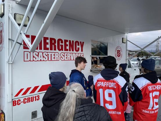―
A medium shot shows a white Salvation Army Emergency Disaster Services truck with the words "EMERGENCY DISASTER SERVICES" in red letters. People are lined up at a window in the truck, where someone is serving food. The truck has a red and white Salvation Army logo, and red stripes with "BC-CRU13" written in the lower corner of the truck. People are wearing winter clothing such as jackets and beanies. Two people are wearing red and white sports jerseys, one with the name "SPAFFORD" and the number "19" and the other with the name "BETCHER" and the number "12". The jerseys also have the word "AVIATION" written on them. In the background, there are other vehicles and buildings.
―