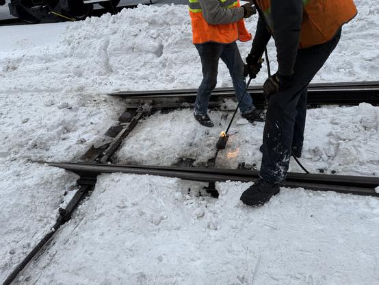 A rail switch with two people standing over it, one with a large blowtorch melting ice off the mechanism