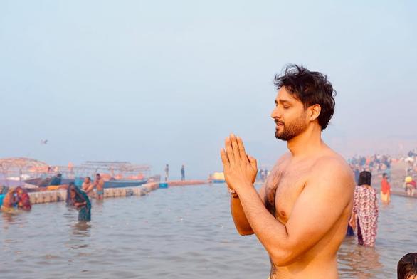 Sourabh Raaj Jain prepares to immerse himself in the Triveni Sangam, where the sacred Ganga, Saraswati and Yamuna rivers converge, at Prayagraj during the Maha Kumbh Mela. He is visible from the waist up, unclothed, seen from his left side as he stands in the middle of the water, with others bathing partially clothed further away in the water. His tattoo on the left side of his chest is partly visible, obscured by the angle and his arms, and there is a plain gold stud in his ear. His eyes are closed, his mouth slightly open as though reciting a mantra, and his hands are raised in front of him, palms together, in an anjali mudra prayer gesture. The photo was shared by Sourabh in a post on Instagram on Feb. 1, 2025.