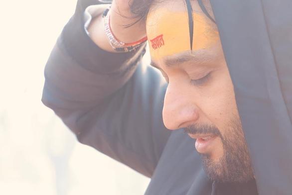 A close up of Sourabh Raaj Jain's face, seen from the side, as he prepares to immerse himself in the Triveni Sangam, where the sacred Ganga, Saraswati and Yamuna rivers converge, at Prayagraj during the Maha Kumbh Mela. Tumeric paste has been applied across his forehead, with characters in Devanagari written in red kumkum. His eyes are closed and his arms are raised, his hands placed on or above his head, out of frame. He is wearing a dark blue jacket, and bands on his right wrist. The photo was shared by Sourabh in a post on Instagram on Feb. 1, 2025.