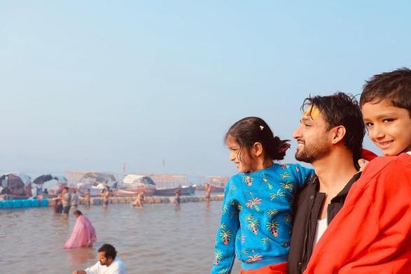 Sourabh Raaj Jain sits with Hrishika and Hrishivh on the banks of the Triveni Sangam, where the sacred Ganga, Saraswati and Yamuna rivers converge, at Prayagraj during the Maha Kumbh Mela. Hrishika wears a turqoise blue pullover with cartoon palm tree motifs, while Hrishivh wears a bright orange fleece sweater. Sourabh wears a white vest and a dark blue jacket. Tumeric paste and lettering in red kumkum have been applied to his forehead, and there is a plain gold stud in his ear. In the bottom left of the frame, his make-up artist and assistant Chandan Chaurasiya can be partially seen, in a white t-shirt bathing in the water. The photo was shared by Sourabh in a post on Instagram on Feb. 1, 2025.