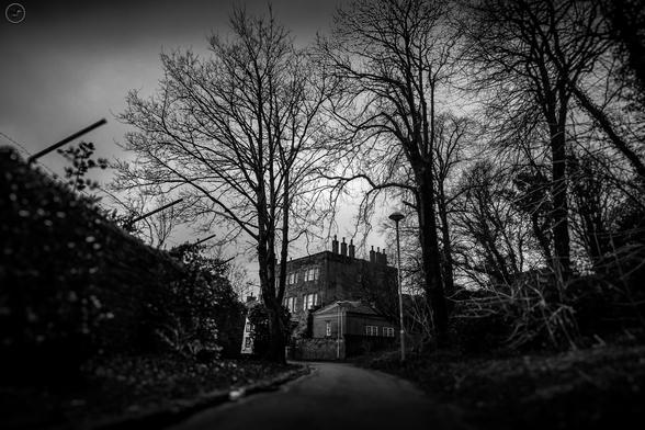 Monochrome shot of period building at end of park path, old wall to left of frame and trees to right. Focused on the building in background