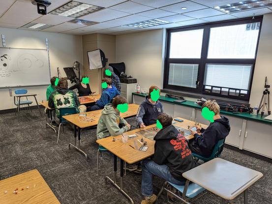 Nine high school students sitting at two tables in a classroom. They are playing Warhammer 40,000 with Space Marines and Necrons.