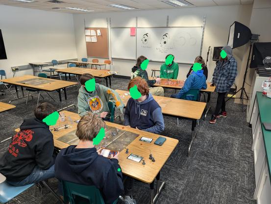 Nine high school students sitting at two tables in a classroom. They are playing Warhammer 40,000 with Space Marines and Necrons.
