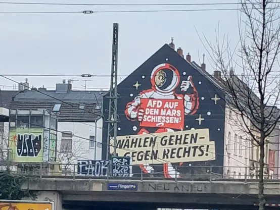 Hauswand nahe Flingern S mit Fassadengestaltung: Ein Astronaut hält ein rotes Schild mit weißer Schrift "AFD AUF DEN MARS SCHIESSEN" und zeigt mit der anderen Hand 👍. Unterhalb ist ein Banner beschriftet mit "WÄHLEN GEHEN - GEGEN RECHTS!"