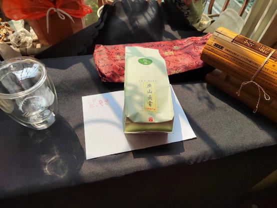 The tea and the tools for the photo shoot laid out in preparation.  A window sill is covered by a black tablecloth with sun shining from above.  To the left is a glass double-walled cup with a tea leaf basket ready to do the brewing.  To the right is a replica bamboo scroll with The Classic of Tea inscribed on it.  Behind that, closer to the background, is the burgundy brocade bag that holds the scroll.  In between the cup and the scroll is a rectangular piece of paper upon which the bag holding the tea is place.  The paper has some seal stamps on it (which will be detailed in later alt text).  The bag with the tea will too be explored in more detail in later alt text.