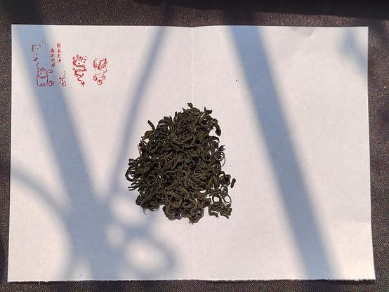 Here a suitable amount of the tea is placed on the fold in the middle of the paper (I fold it so it's easier to put into the cup after I've taken the photo).  This tea is a very dark green in the leaf (one of the ways you identify a cloud and mist tea) and the leaves are carefully rolled lengthwise into almost a little worm shape.  Here the three seal stamps are more clearly visible.  The two right-most ones are a dragon design and a phoenix design which I placed there because I like the emblems.  The leftmost one is an illustration stamp featuring an old-time tea pot on an old-time stove with a couplet I can't decode at the top right (but it's about tea).