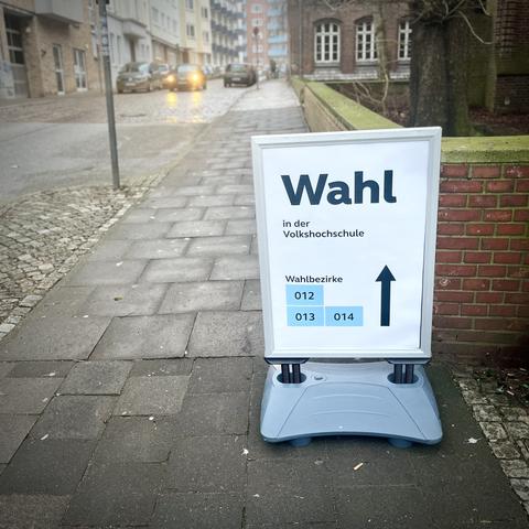 Ein Schild mit dem Hinweis auf einen Wahllokal an einer Volkshochschule mit dem Wort "Wahl" und Wegbeschreibungen zu den Wahlbezirken 012, 013 und 014. Das Schild befindet sich auf einem Bürgersteig an einer Straße.